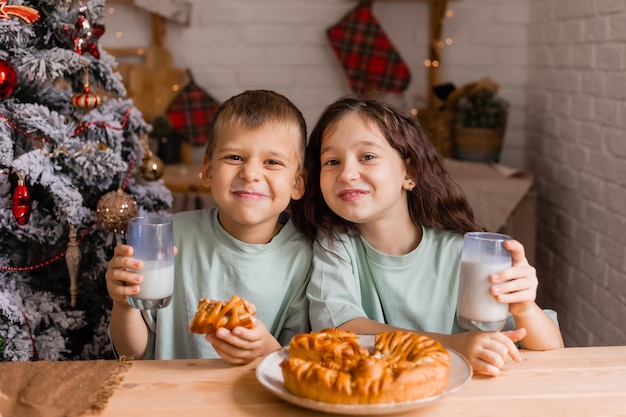 Dwoje małych dzieci, chłopiec i dziewczynka, jedzą ciasto świąteczne z mlekiem rano w domu w kuchni