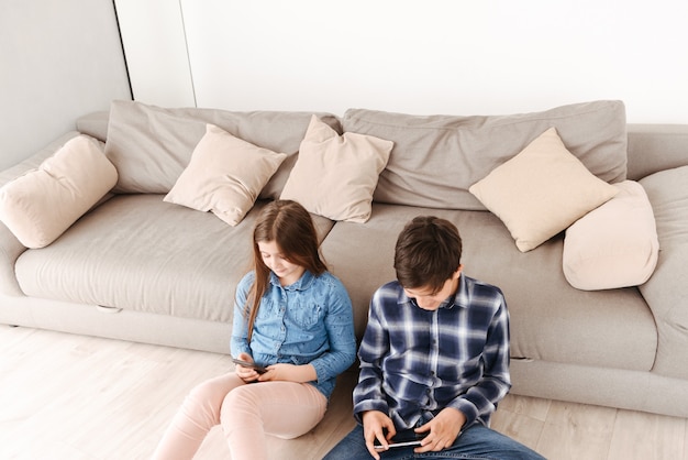 Dwoje Nowoczesnych Dzieci, Dziewczyna I Chłopak, Siedzący Na Podłodze W Pobliżu Sofy W Domu I Oboje Za Pomocą Smartfona