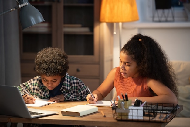 Dwoje słodkie rodzeństwo pochodzenia afrykańskiego siedzi przy stole w domu i robi notatki w zeszytach przed laptopem podczas oglądania lekcji online