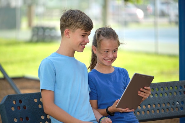 Dwoje szczęśliwych dzieci, dziewczyna i chłopak, patrząc na ekran tabletu cyfrowego, czytając, studiując lub grając, siedząc na ławce na świeżym powietrzu w słoneczny letni dzień