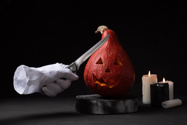 Zdjęcie dynia z rzeźbioną twarzą i niewidzialną ręką w białej rękawiczce z nożem, płonące świeczki na czarnym tle, przygotowanie jack-o-lantern na halloween, zbliżenie.