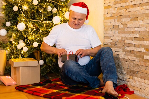 Dziadek siedzi przy drzewie Cristmas w kapeluszu Świętego Mikołaja w nocy w domu.