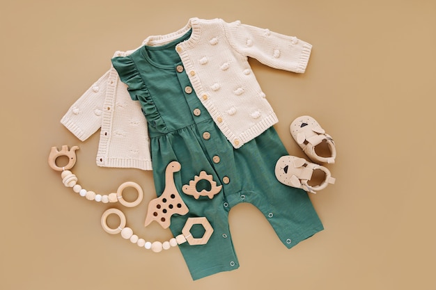 Dzianinowy sweter z zielonym pajacykiem, drewnianymi zabawkami i bucikami dla niemowląt. Zestaw ubranka i akcesoria dla dziewczynki na beżowym tle. Moda noworodka. Płaski układanie, widok z góry