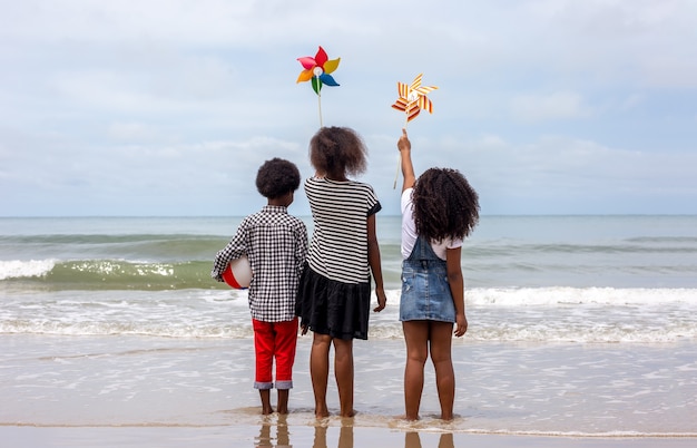 Dzieci bawiące się bieganie po piasku na plaży, grupa dzieci trzymających się za ręce z rzędu na plaży latem, widok z tyłu na morze i błękitne niebo