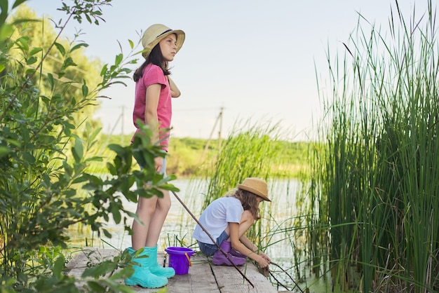 Dzieci dwie dziewczyny razem bawić się wodą na jeziorze na drewnianym molo w trzcinach. Dzieci z wiaderkiem do zbierania patyczków z alg i ślimaków wodnych. Natura, wypoczynek, przyjaźń, koncepcja wakacji letnich