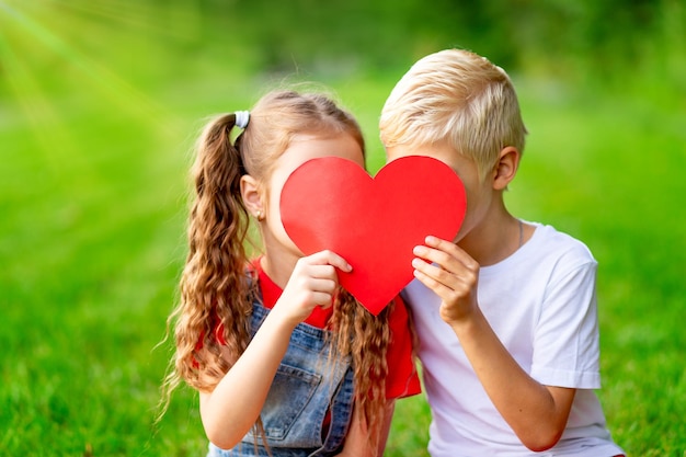 Dzieci dziewczyna i blond chłopiec całują się za dużym czerwonym sercem latem na trawniku na zielonej trawie koncepcja walentynkowej przestrzeni wakacyjnej na tekst
