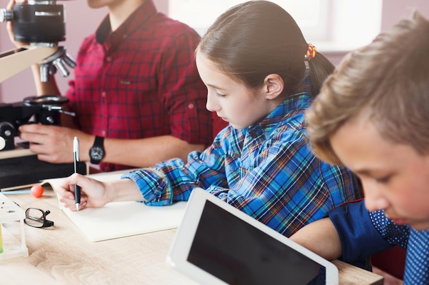 Dzieci I Nauka. Zbliżenie: Uczniowie Wykonujący Szkolny Projekt Klasowy W Laboratorium, Używający Tabletu I Mikroskopu Oraz Pisania Wyników. Wczesny Rozwój, Innowacja, Edukacja Macierzysta, E-learning