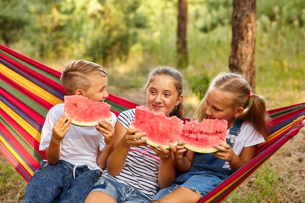 Dzieci jedzą arbuzy i żartują siedząc na kolorowym hamaku na świeżym powietrzu