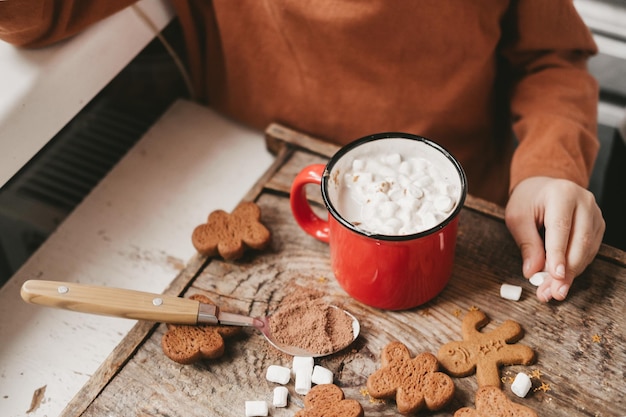 Dzieci piją kakao z marshmallows i świątecznym piernikiem na drewnianym tle. Świąteczne słodycze z kakao i piankami dla dzieci. Zimowe wakacje w domu