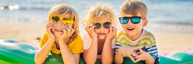 Zdjęcie dzieci siedzą na dmuchanym materacu w okularach przeciwsłonecznych na tle morza i bawią się banerem w długim formacie