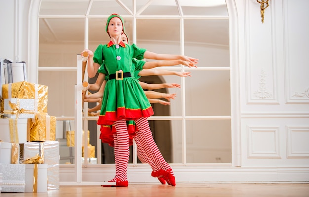Dzieci ubrane w kostiumy bożonarodzeniowych elfów ćwiczą przy barze baletowym przed lustrem w przestronnym białym studio.