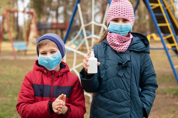 Dzieci w maskach medycznych używających dezynfekcji rąk podczas zabawy na placu zabaw na świeżym powietrzu. Życie podczas kwarantanny koronawirusa