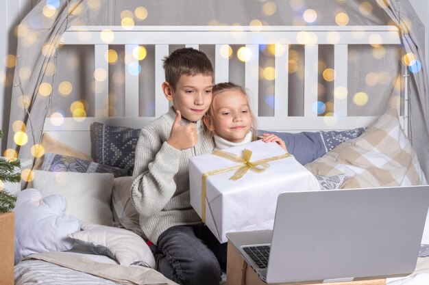 Dzieci w wieku przedszkolnym trzymające pudełko na prezent rozmawiające za pośrednictwem łącza wideo za pośrednictwem laptopa i machające ręką na powitanie krewnych lub przyjaciół w domu