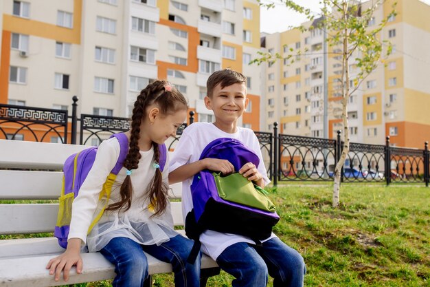 Dzieci w wieku szkolnym chłopiec i dziewczynka siedzą na ławce z jasnymi plecakami i uśmiechają się, patrzą w kamerę