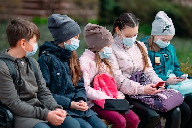 Dzieci w wieku szkolnym w maskach medycznych siedzą na ławce.