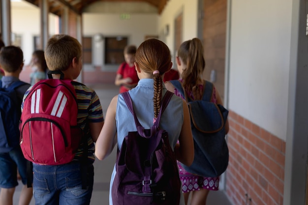 Dzieci ze szkół podstawowych. Widok z tyłu zróżnicowanej grupy uczniów noszących plecaki spacerujących po korytarzu zewnętrznym w szkole podstawowej