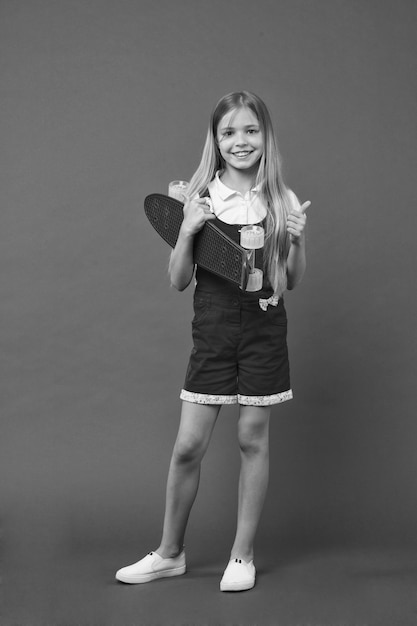 Dzieciak dziewczyna gotowa jeździć na deskorolce Współczesne hobby młodzieżowe Deskorolki znane również jako mini krążownik Dziewczynka szczęśliwa buźka trzyma deskę na fiszkę fioletowe tło Pierwotnie zaprojektowany jako deskorolka dla dziewczynek
