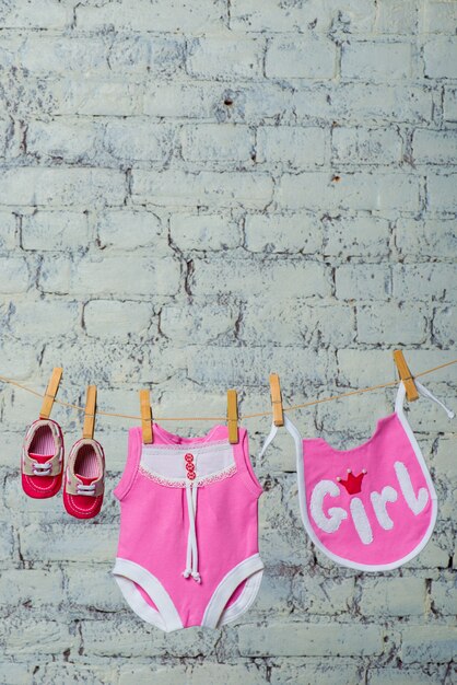 Zdjęcie dziecięcy różowy śliniaczek i czerwone buty wysychają na linie na białej ścianie z cegły