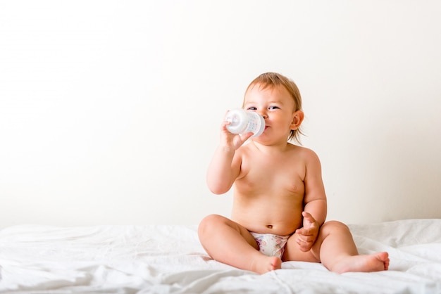 Dziecko berbeć siedzi na białym łóżku, ono uśmiecha się i pije wodę z plastikowej butelki