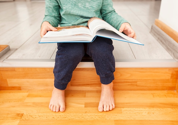 Zdjęcie dziecko czyta papierową książkę siedząc na drewnianych schodach w domu
