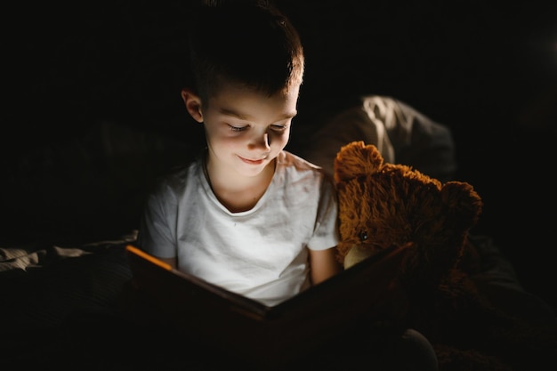 Dziecko Czytanie Książki W łóżku. Dzieci Czytają W Nocy. Mały Chłopiec Z Książkami Bajek W Sypialni. Edukacja Dla Małych Dzieci. Bajka Na Dobranoc Wieczorem. Słodkie Dziecko Pod Kocem W Ciemnym Pokoju Z Lampą.