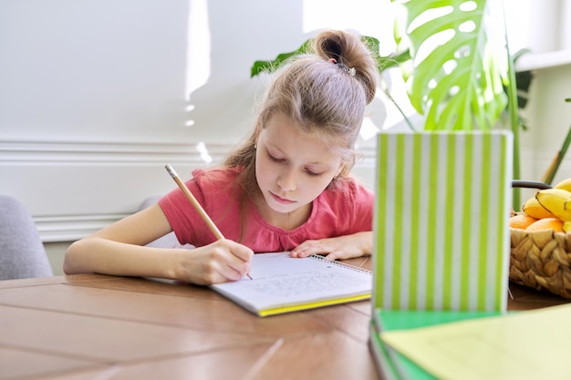 Dziecko dziewczynka studiuje w domu, siedząc przy stole. Uczennica pisze w zeszycie, zagląda do książek, nauki na odległość, nowoczesnej szkoły i edukacji