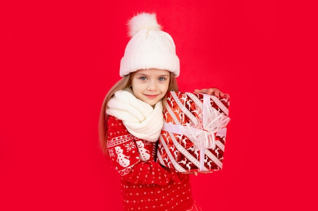 Dziecko dziewczynka w zimowym kapeluszu i swetrze z prezentami na czerwonym monochromatycznym na białym tle raduje się i uśmiecha się koncepcją nowego roku i świątecznego miejsca na tekst