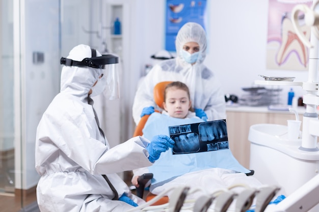 Dziecko i matka w garniturze ppe słuchający dentysta mówi o procedurze dentystycznej. Stomatolog w kombinezonie ochronnym na koronawirusa jako środek ostrożności trzymający prześwietlenie zębów dziecka podczas konsultacji.