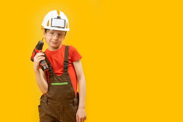 Dziecko jako budowniczy Chłopiec w białym hełmie okulary ochronne nosi kombinezon roboczy i trzyma wiertło Odizolowane na żółtym tle