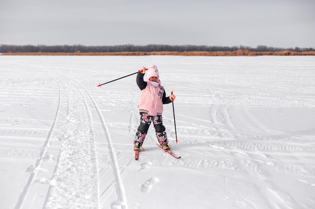 Dziecko jeździ na nartach w śniegu Szczęśliwa dziewczyna w ciepłym dresie uczy się jeździć na nartach na zamarzniętym brzegu rzeki i macha ręką zimowy krajobraz śnieżny tło