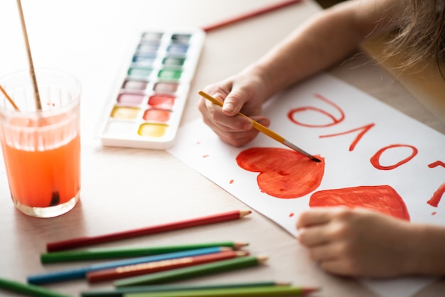 Zdjęcie dziecko maluje akwarelami akwarela czerwone serce na arkuszu krajobrazu na drewnianym stole