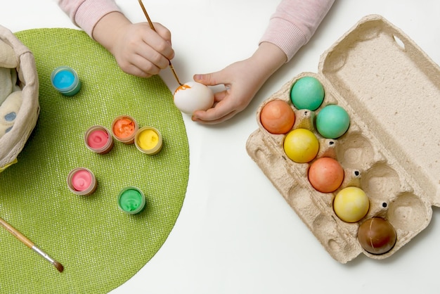 Dziecko maluje jajka kolorowymi farbami i wkłada je do koszyka z zawartością wielkanocną