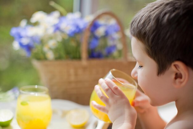 Dziecko pije lemoniadę z owocami cytrusowymi i miętową koncepcją letnich napojów Szklanki z lemoniadą z miętowymi cytrynami limonki i pomarańczą na białej drewnianej tacy w pobliżu słomianej torby z polnymi kwiatami