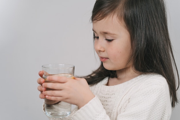 Dziecko pije wodę ze szklanki. Dziewczyna o ciemnych włosach trzyma szklankę wody. Brunetka prowadzi zdrowy tryb życia