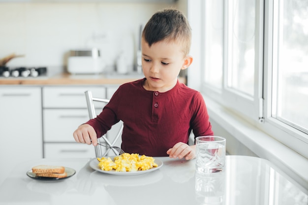 Dziecko po południu w białej kuchni w bordowym swetrze zjada omlet