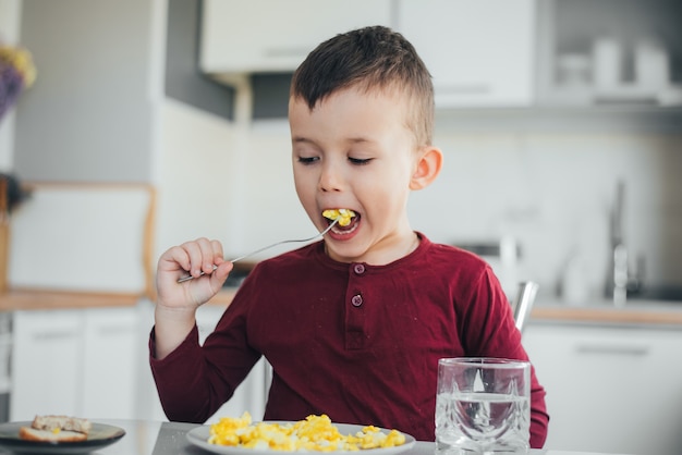 Dziecko po południu w białej kuchni w bordowym swetrze zjada omlet