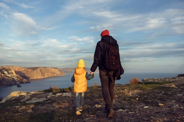 Dziecko-podróżnik z ojcem spaceruje po klifie nad morzem w kurtkach, plecami do zachodu słońca