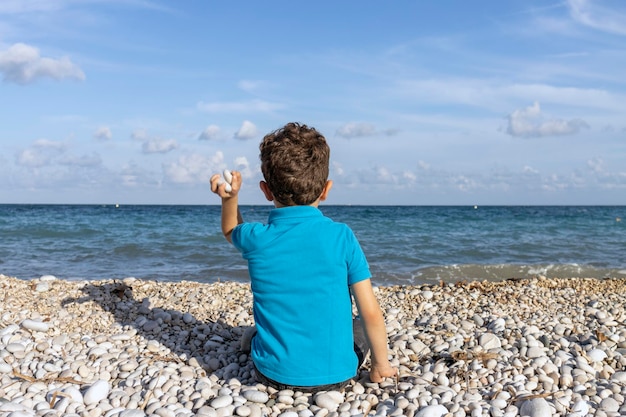 Dziecko siedzące na brzegu plaży rzucające kamieniami do wody widziane od tyłu