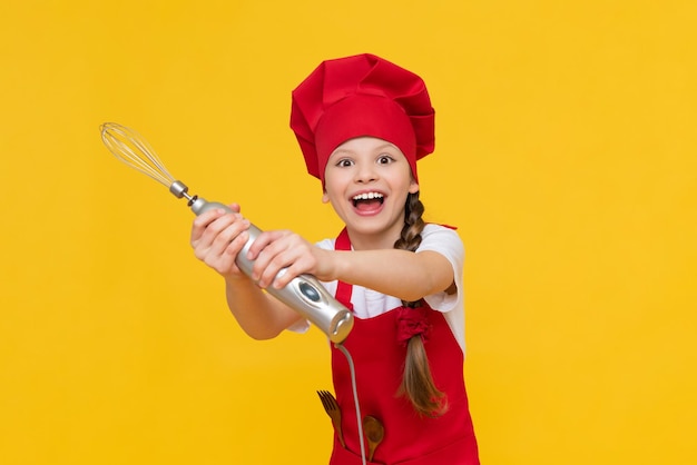 Dziecko szefa kuchni trzyma blender do ubijania Szczęśliwa mała dziewczynka w czerwonym kapeluszu i fartuchu Gotowanie potraw Żółte odosobnione tło