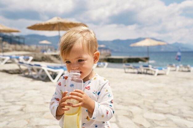 Dziecko trzyma butelkę wody, stojąc na brukowanej plaży z leżakami