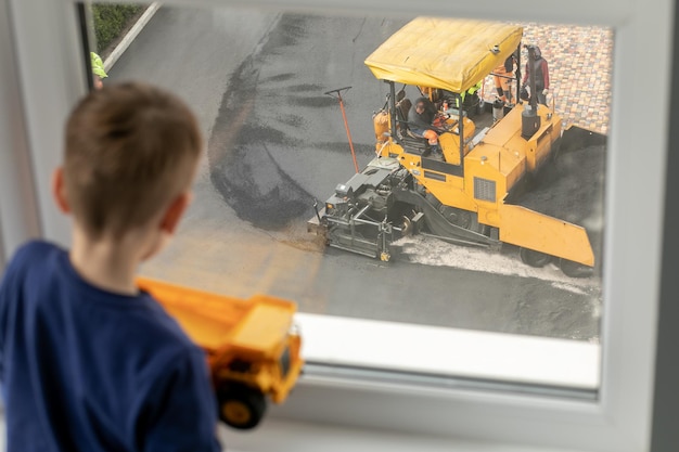 Dziecko trzyma samochodzik, wygląda przez okno, w którym pracują konstruktorzy okien, marzenie ma zostać budowniczym
