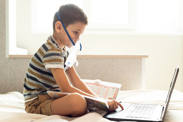 Dziecko uczy się lekcji w domu w masce tlenowej z nibulizatorem, laptopem i książką