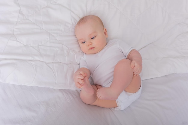 dziecko w białym body leży na plecach w białym łóżku dzieciak bawi się palcami u nóg