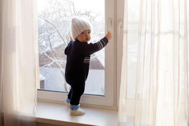 Dziecko w ciepłym niebieskim kombinezonie na oknie w zimowej białej czapce