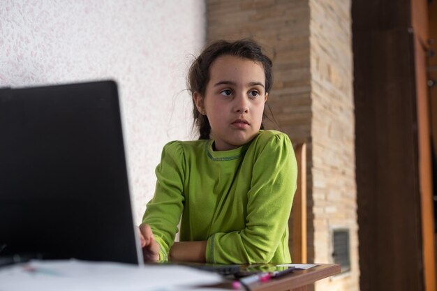 Dziecko w Internecie. Mała dziewczynka korzysta z czatu wideo na laptopie, aby komunikować się z nauką, siedząc przy stole w domu.