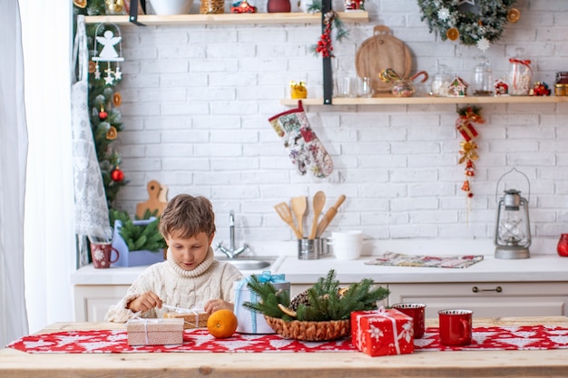 Dziecko w kuchni w Wigilię wczesnym rankiem rozpakowuje prezent od Mikołaja. szczęśliwy chłopiec na kuchennym stole z pudełkami. Wesołych Świąt i Wesołych Świąt!