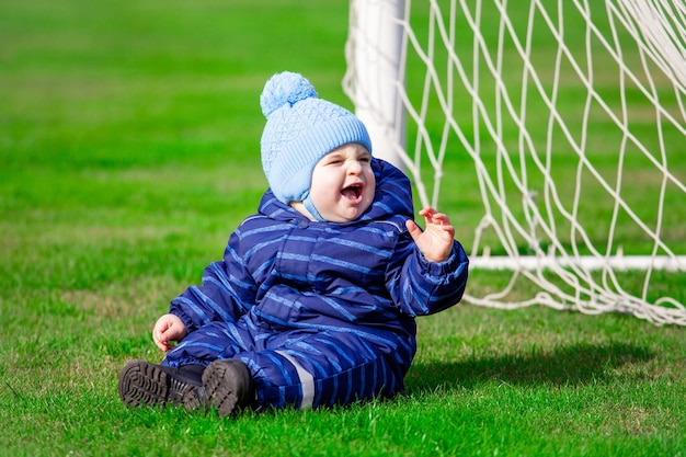 Dziecko w niebieskim kombinezonie siedzi na boisku przy bramce