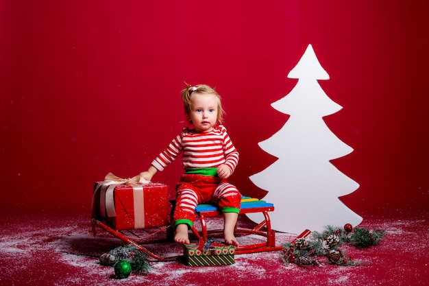 Dziecko W Piżamie I Czapce Mikołaja łapie śnieg Siedząc Na Sankach Z Pudełkiem Prezentowym I Dużą Białą Choinką Na Czerwonym Tle