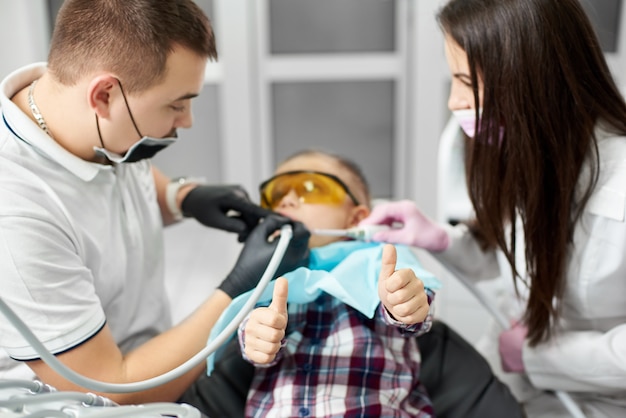 Zdjęcie dziecko w pomarańczowych okularach dentystycznych pokazuje kciuki do góry, podczas gdy dentysta ma na sobie niebieską koszulkę i czarne rękawiczki z asystentem brunetki.