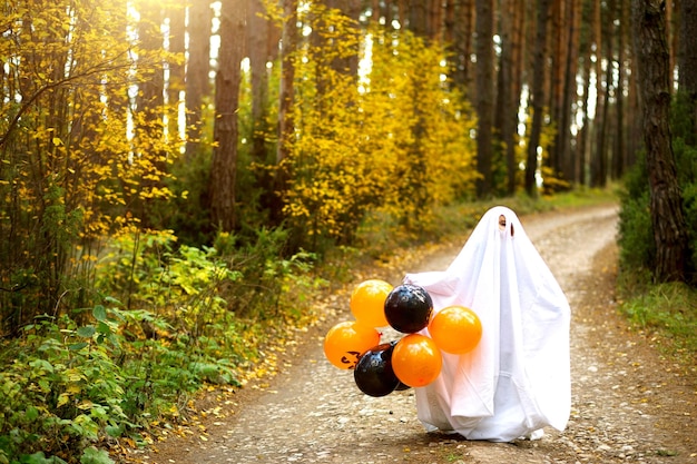 Dziecko w prześcieradłach z rozcięciami jak kostium ducha w jesiennym lesie z pomarańczowymi i czarnymi kulkami przeraża Halloween Party